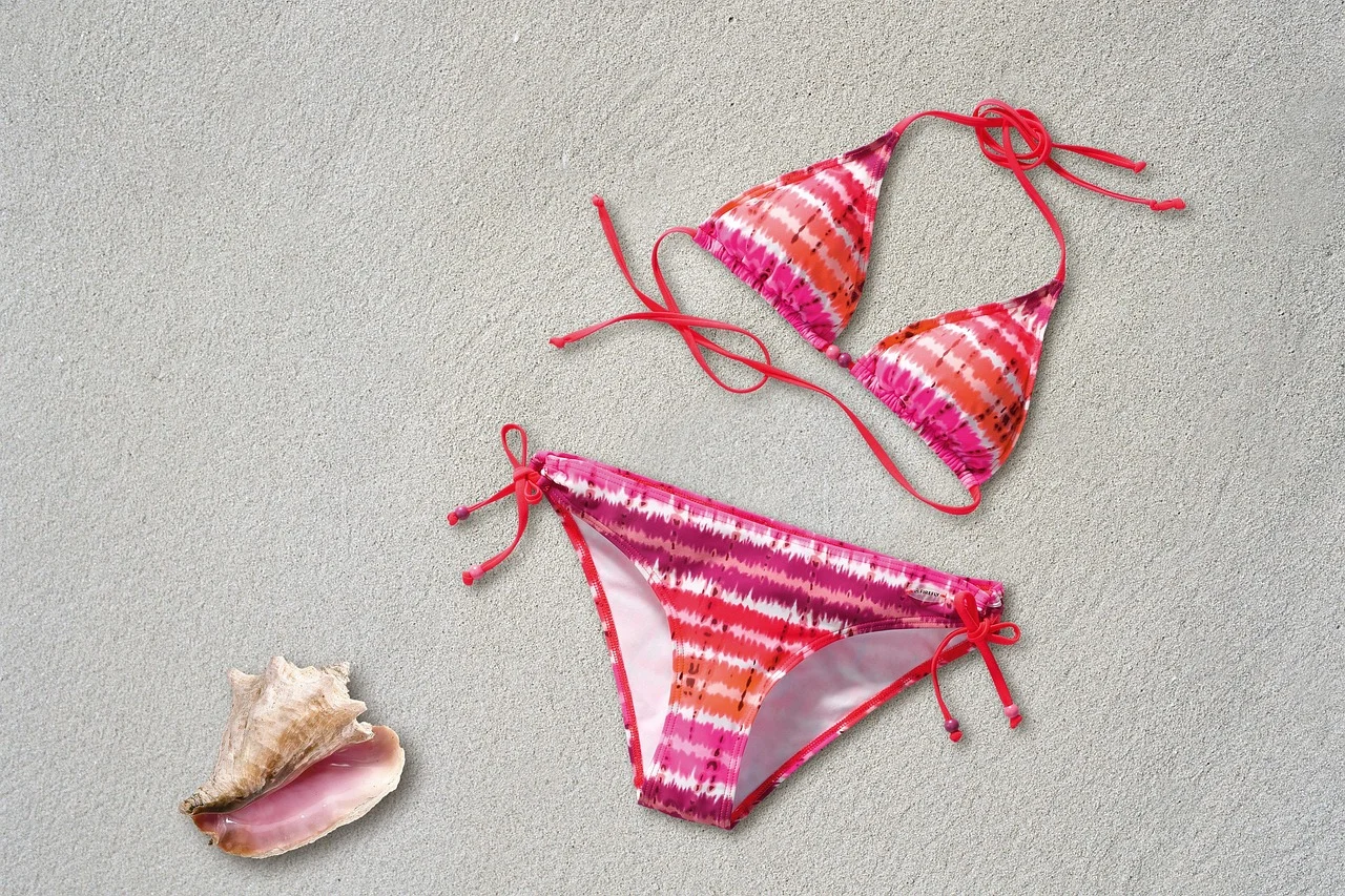 Bikini in beach với họa tiết tie-dye hồng cam nằm trên cát trắng bên vỏ ốc biển